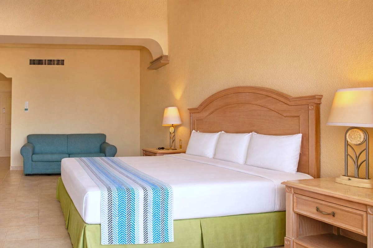 A hotel room with a bed, a green skirted bedspread, a blue and white striped blanket, a wooden headboard, a nightstand with a lamp, a green sofa, and a wall-mounted air vent.