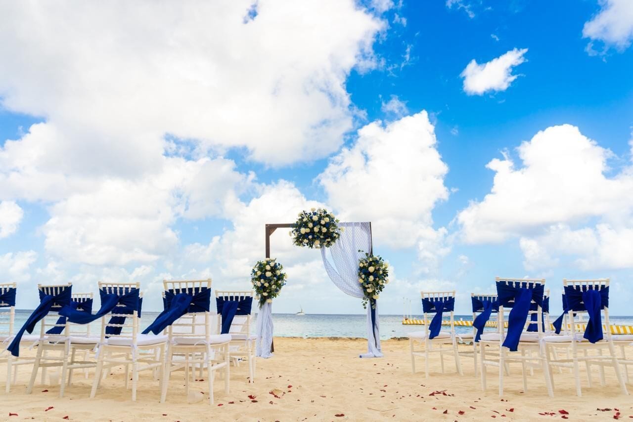 A beach wedding setup with white chairs, blue drapes, and floral decorations.