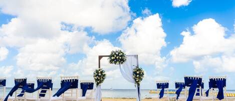 Decoração de casamento na praia, com cadeiras brancas, cortinas azuis e detalhes florais.