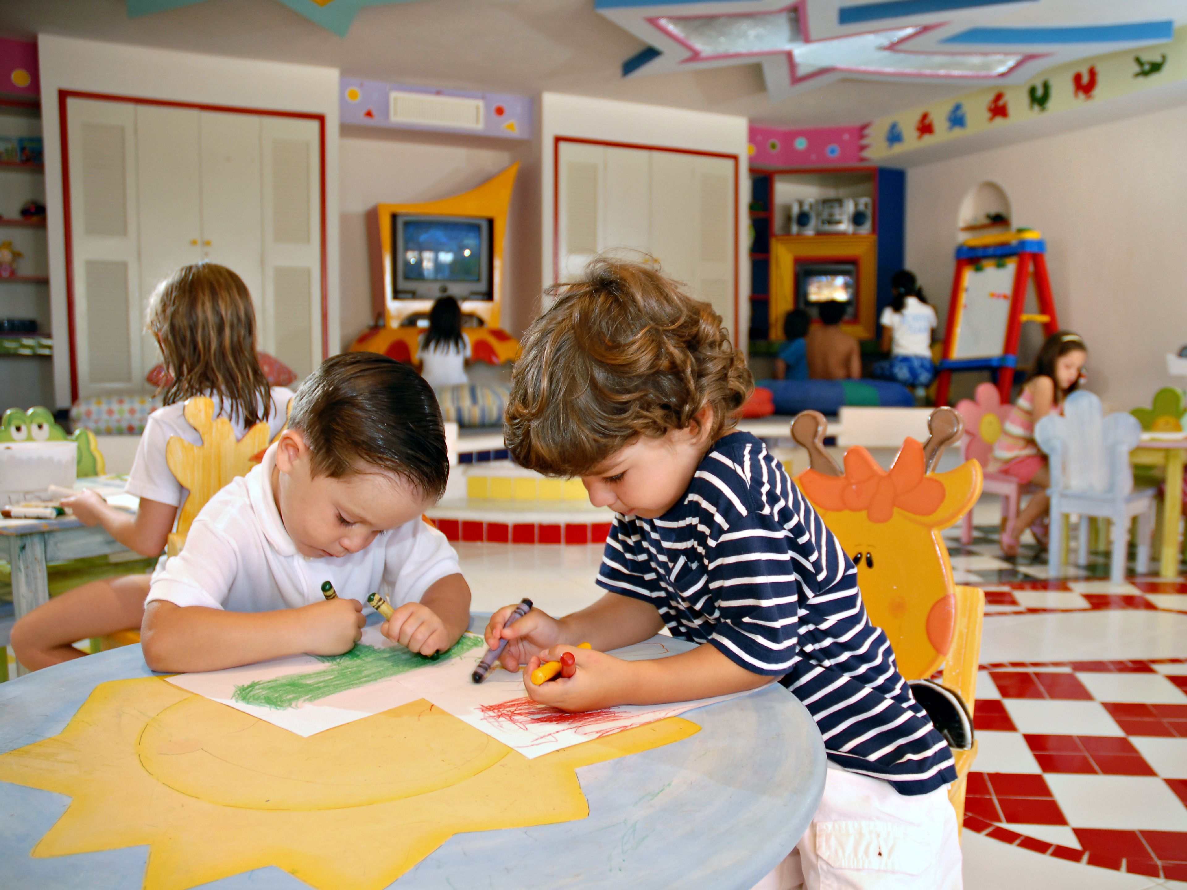 Kinder zeichnen an einem Tisch in einem farbenfrohen Raum mit Lehrmaterialien und Dekorationen.