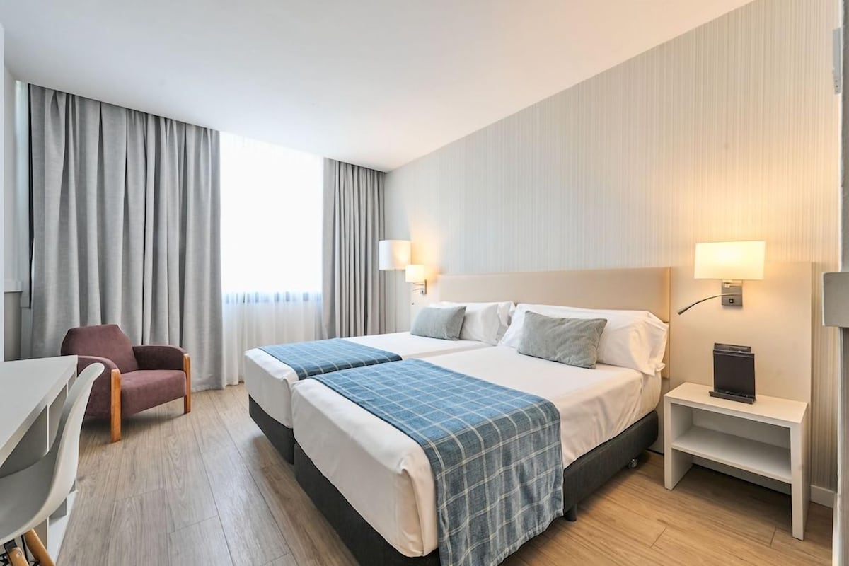 Una habitación de hotel con dos camas, un escritorio, una silla y una mesita de noche con lámpara.