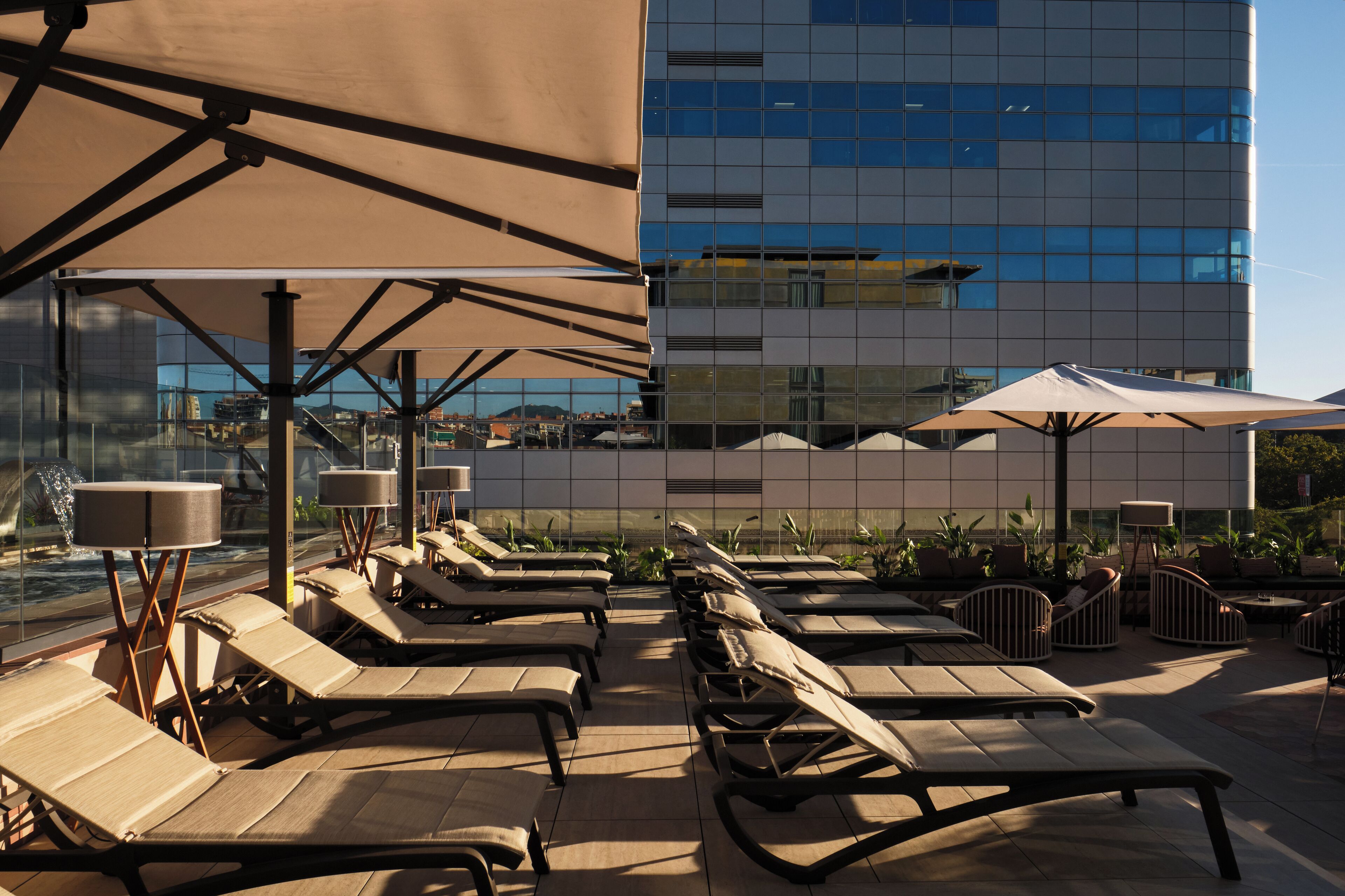 Zona de lounge en la azotea con tumbonas y sombrillas, con vista a un edificio moderno con ventanas reflectantes.