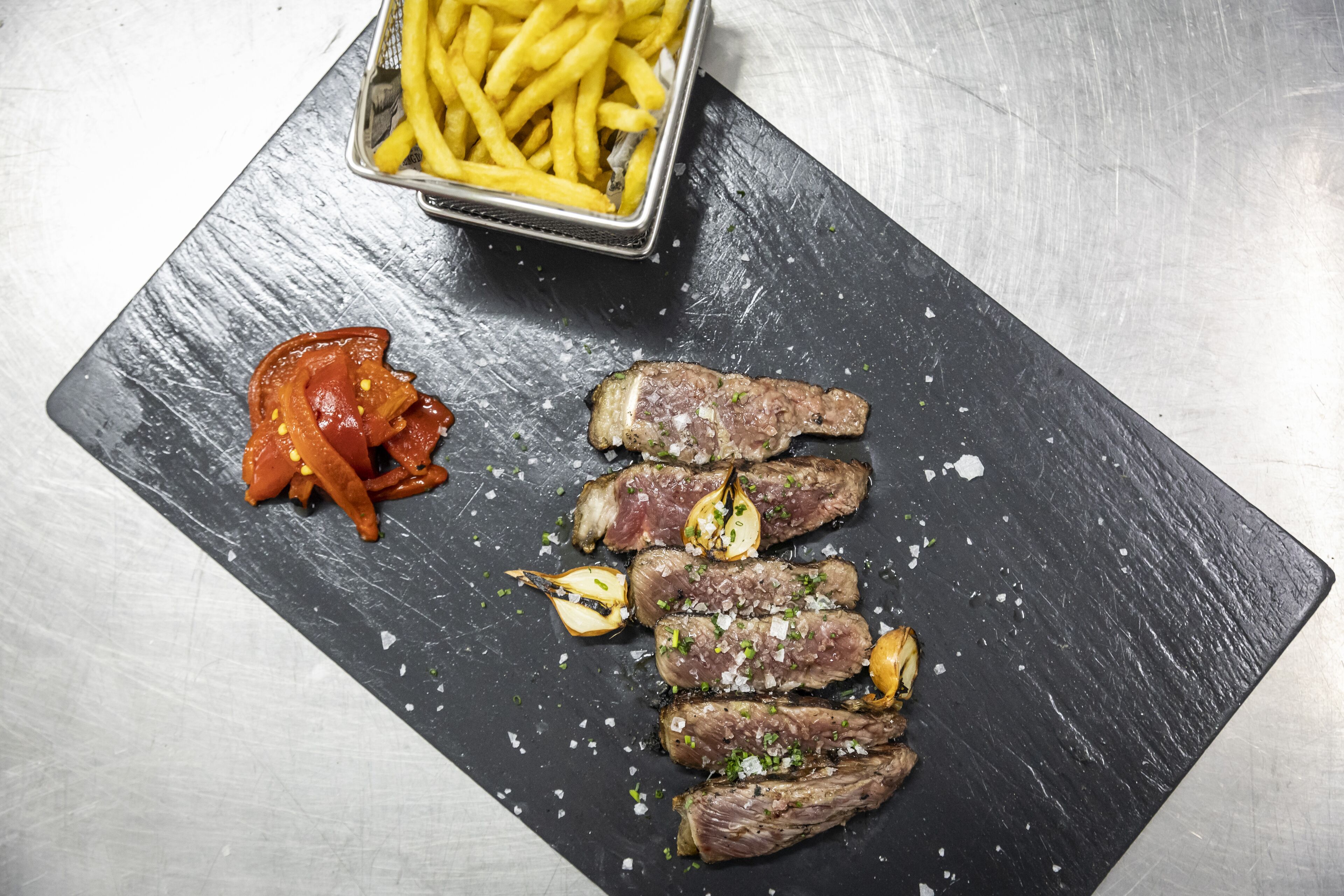 Una pizarra de pizarra negra con carne en rodajas, ajo y un recipiente con papas fritas.