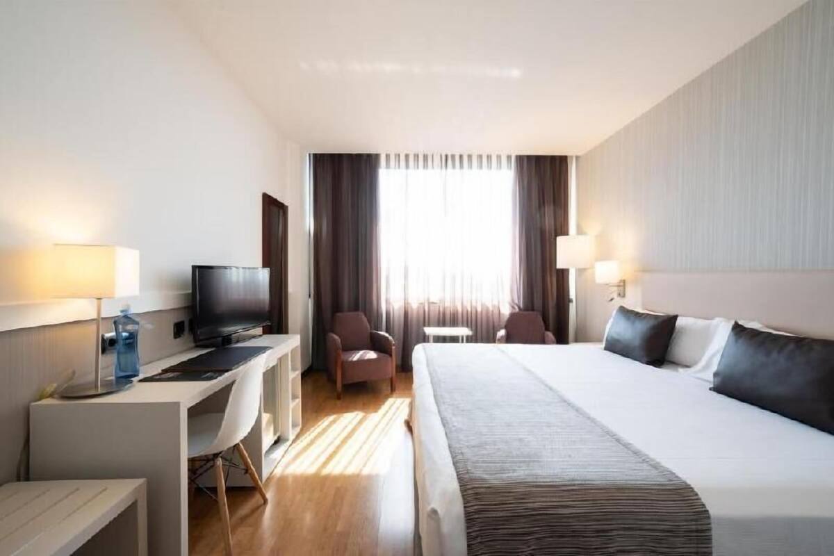 Een hotelkamer met een bed, bureau, televisie, twee stoelen en een raam met gordijnen.