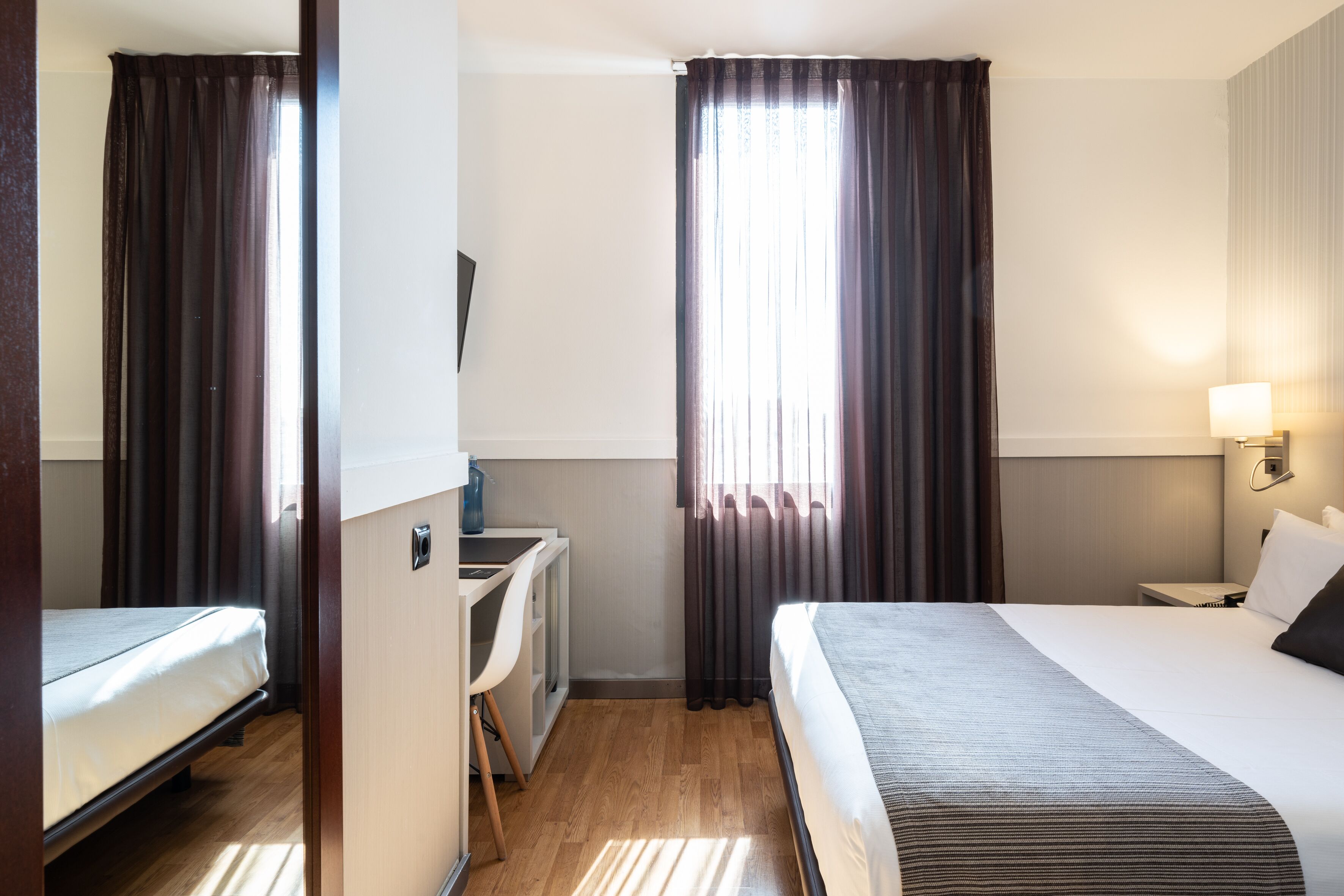 A hotel room with a bed, desk, and chair, a TV, and a window with curtains.