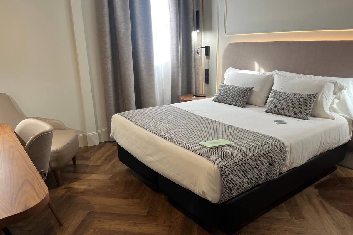 A hotel room with a large bed, a chair, a wooden table, and a trash bin.
