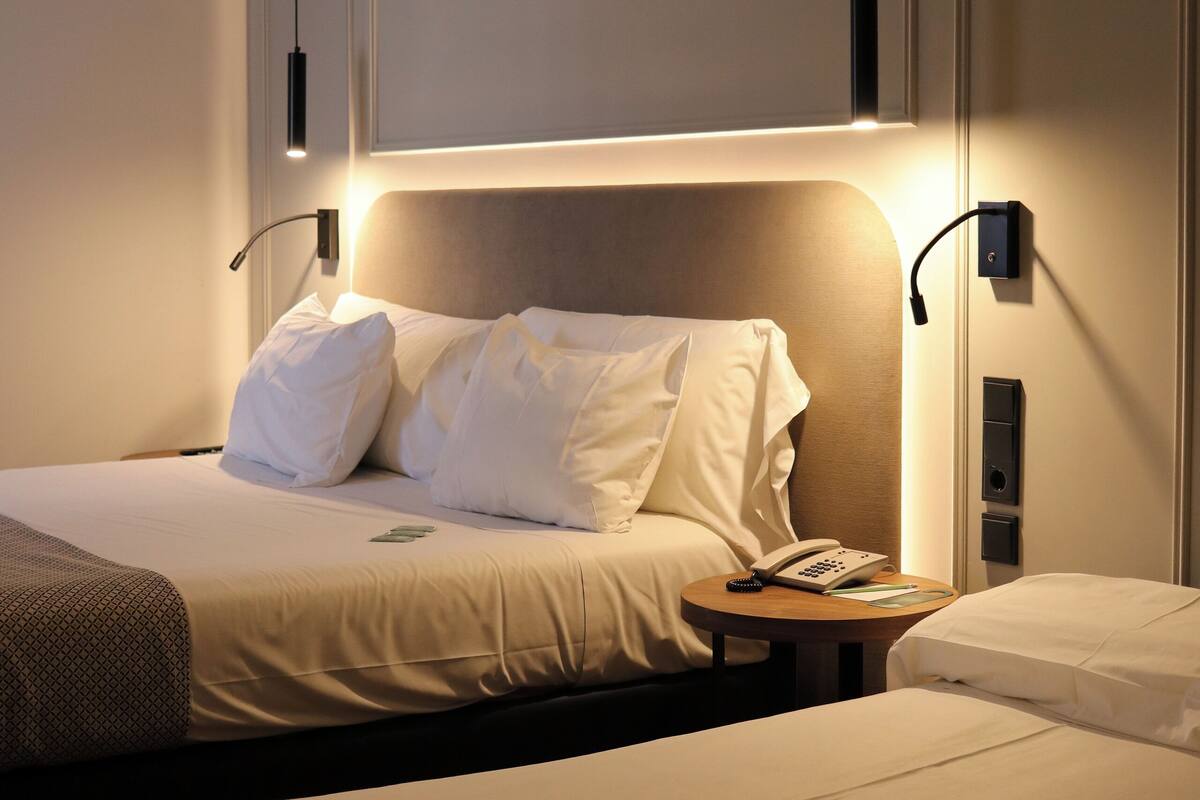 A hotel room with a bed, bedside table, and a small desk with a lamp.