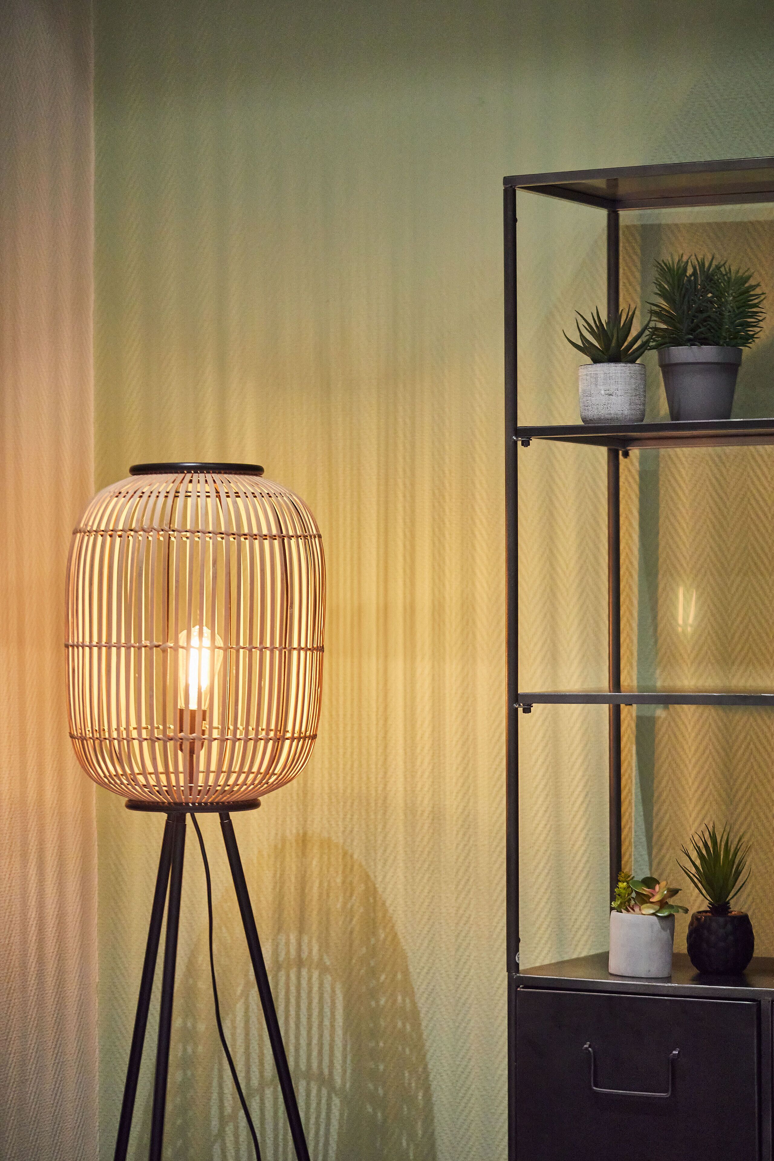 Una lampada da terra con un design cilindrico e scanalato, con una singola lampadina, posizionata accanto a un mobile scaffale in metallo nero con due piante in vaso sullo scaffale superiore.