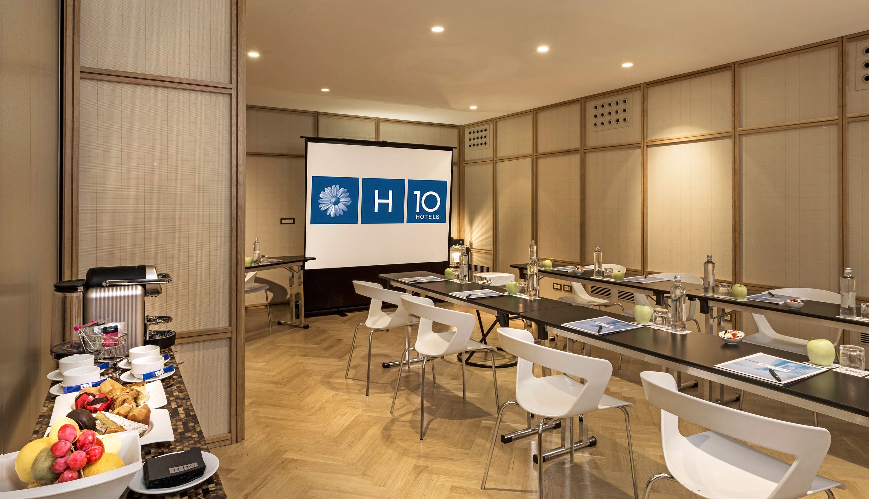 A conference room with a long table, white chairs, a projector screen, and a buffet table with various food items.