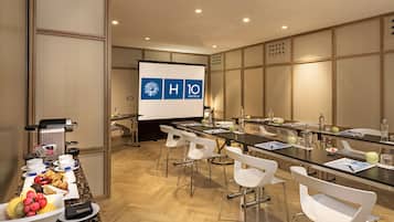 Un salón de conferencias con una larga mesa, sillas blancas, una pantalla de proyector y una mesa de buffet con diversos alimentos.