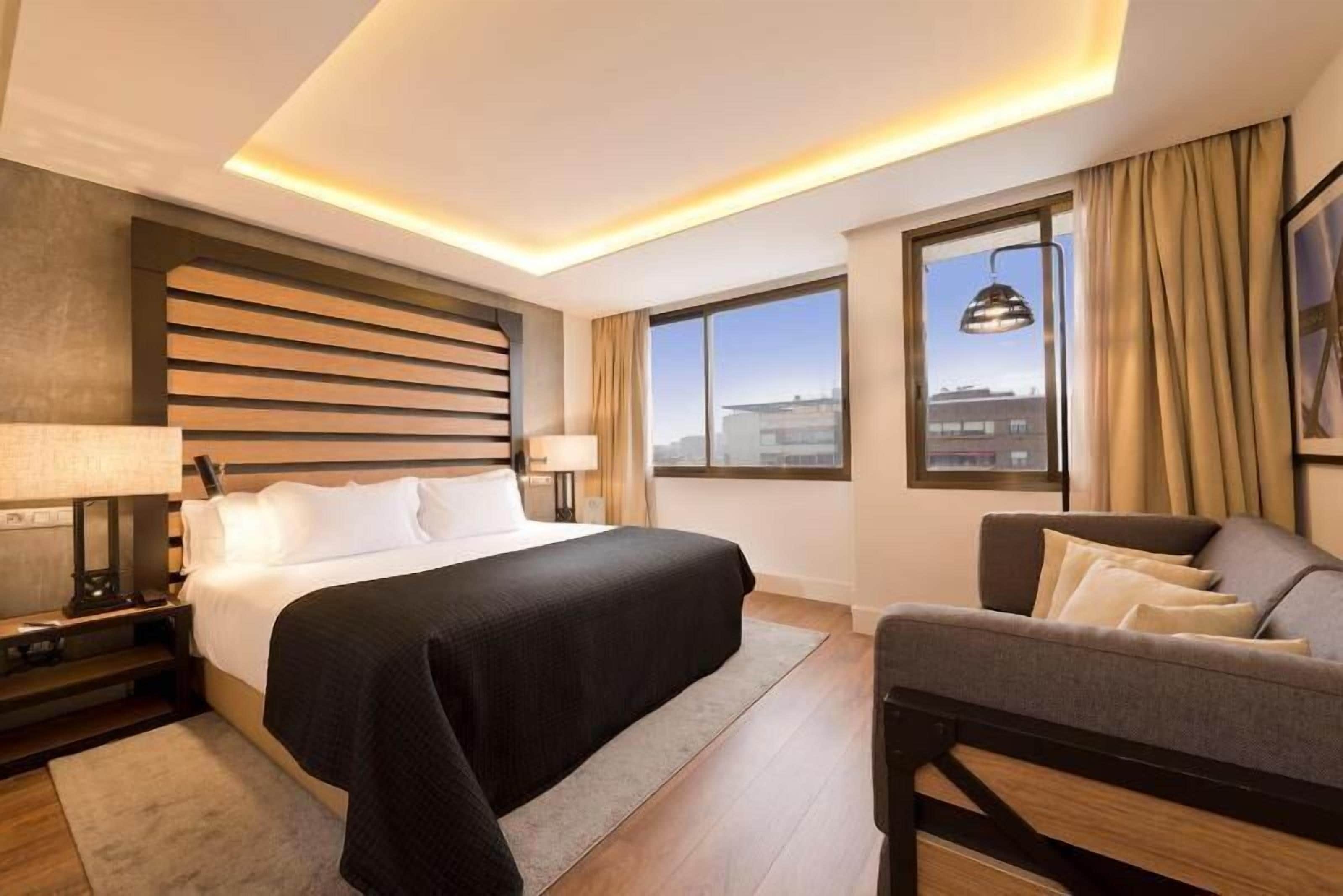 Ett hotellrum med en säng, ett skrivbord, en stol, ett stort fönster och utsikt över staden.
