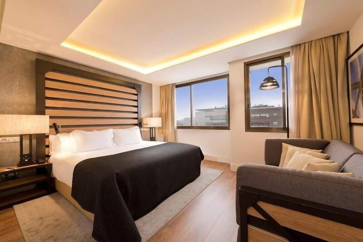 A hotel room with a bed, a desk, a chair, a large window, and a view of the city.