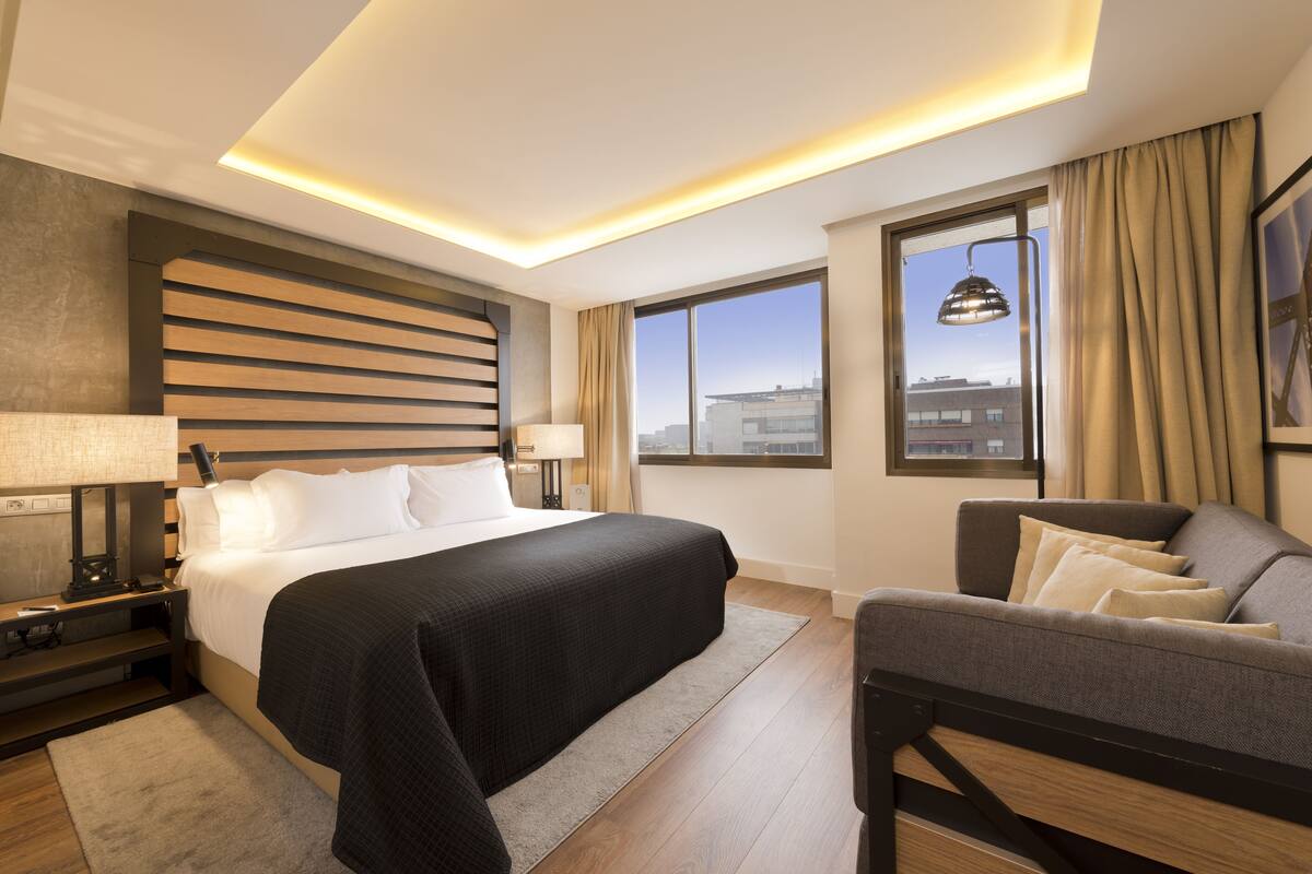 Une chambre d’hôtel moderne avec un grand lit, un canapé, un bureau avec une lampe et une vue sur la ville par la fenêtre.