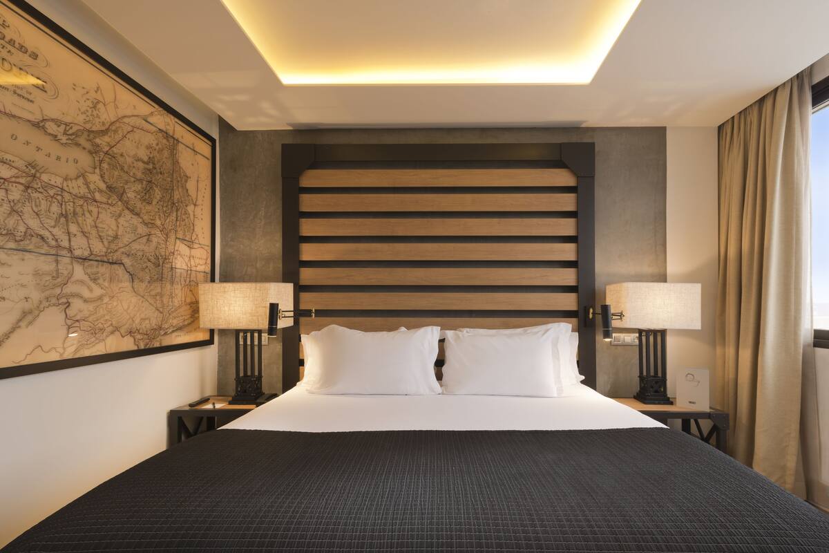 Un lit bien fait, recouvert d’une couverture sombre, deux lampes de chevet, une grande carte encadrée accrochée au mur et une tête de lit moderne.
