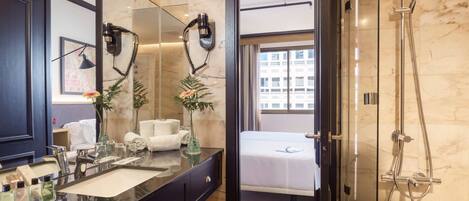 A modern bathroom with a large mirror, a vanity with a sink, a shower area, and a bed visible through an open door.