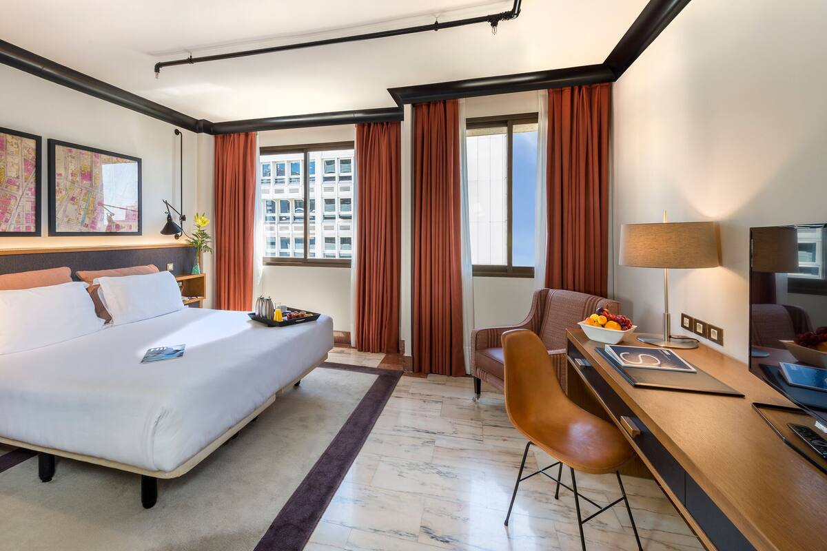 Une chambre d’hôtel avec un grand lit, un bureau avec une chaise, une lampe de bureau, un panier de fruits et une fenêtre avec des rideaux.