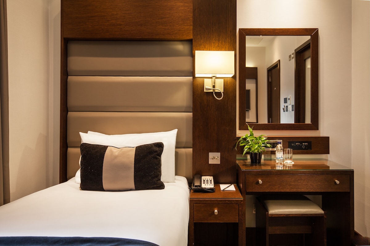 A hotel room with a bed, bedside table, lamp, mirror, and a small desk with a plant.