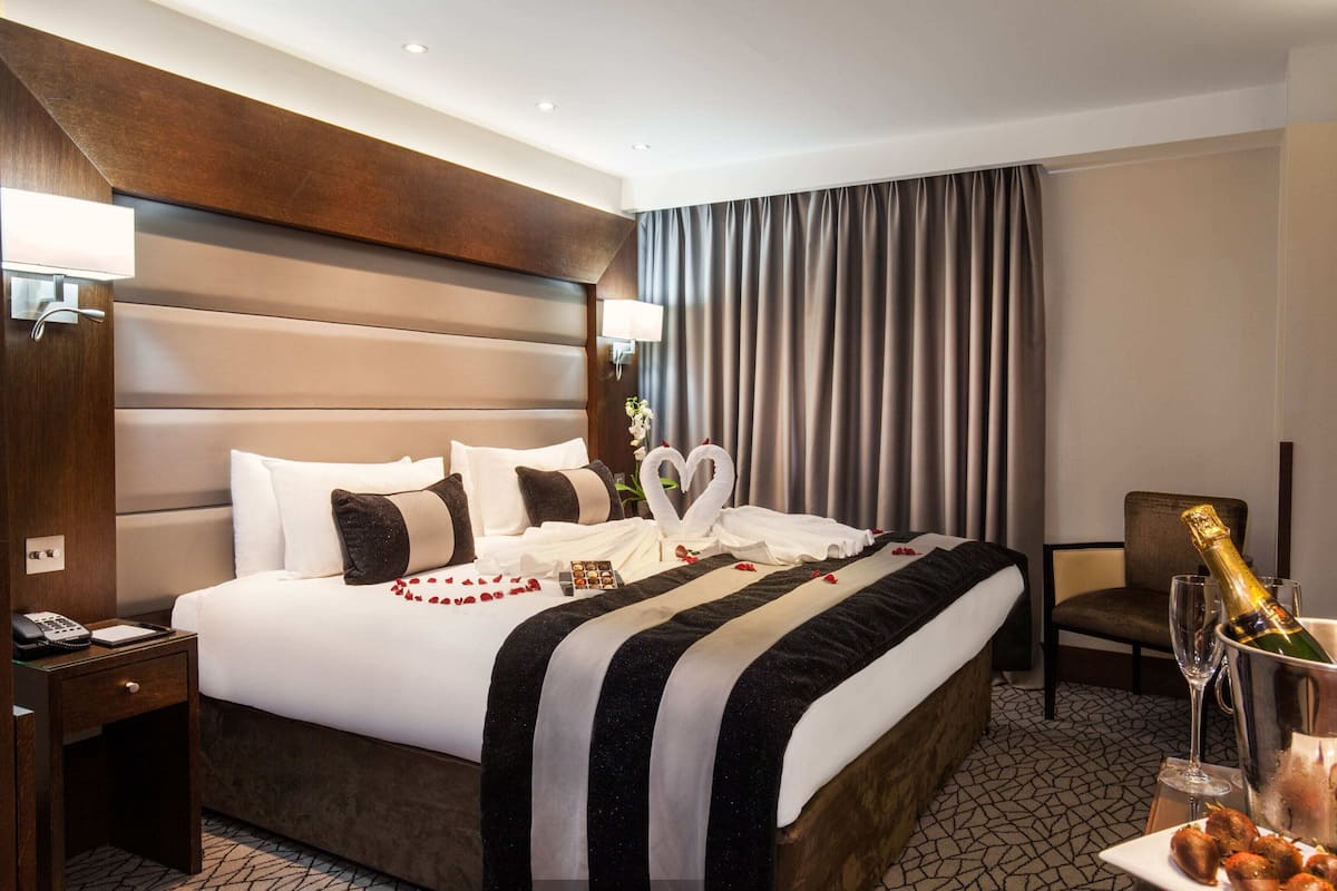 A hotel room with a large bed, a nightstand with a phone, a chair, a bottle of champagne, and a plate of snacks.