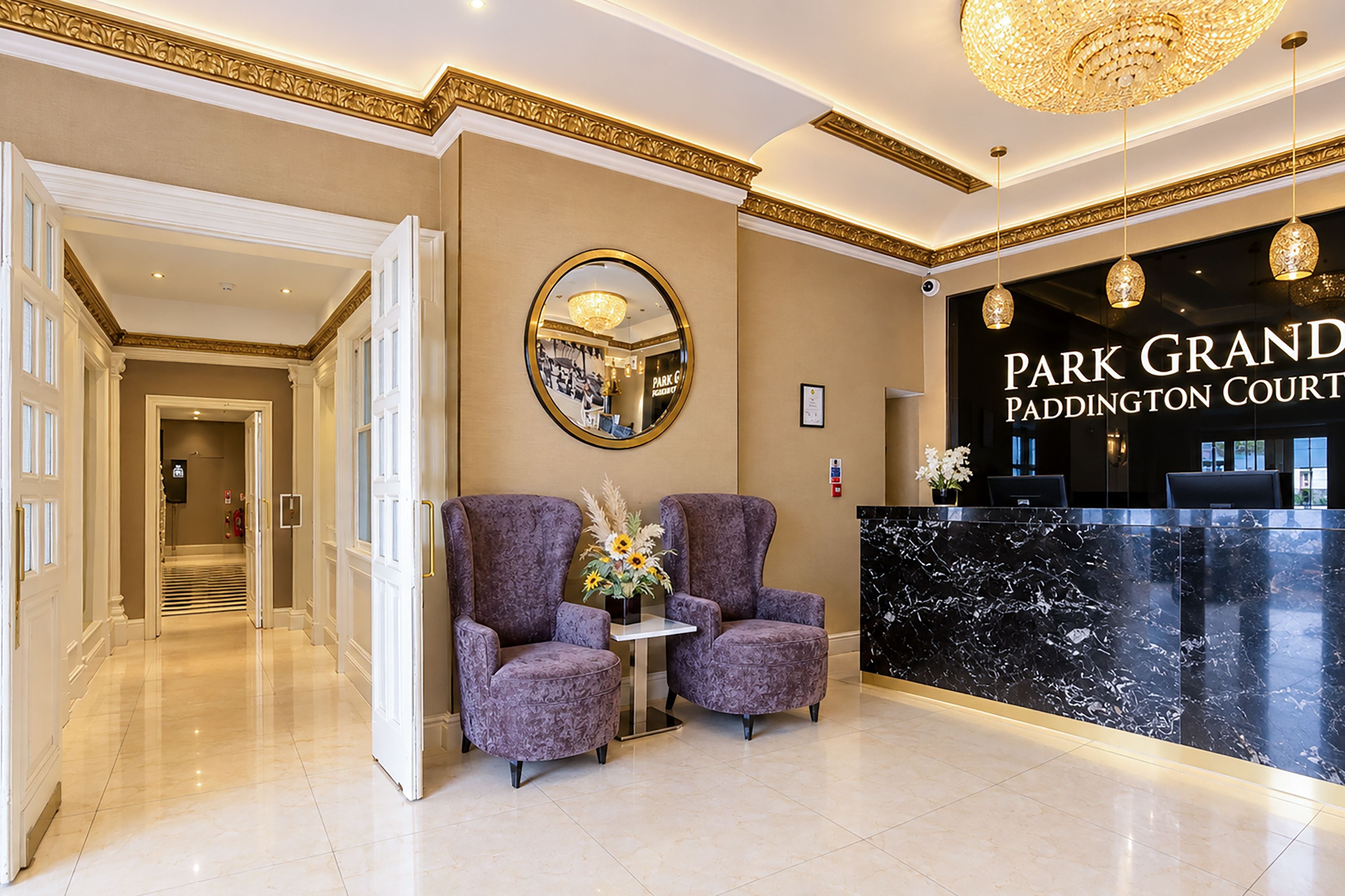 A hotel lobby with a reception desk, two armchairs, a mirror, and a chandelier.