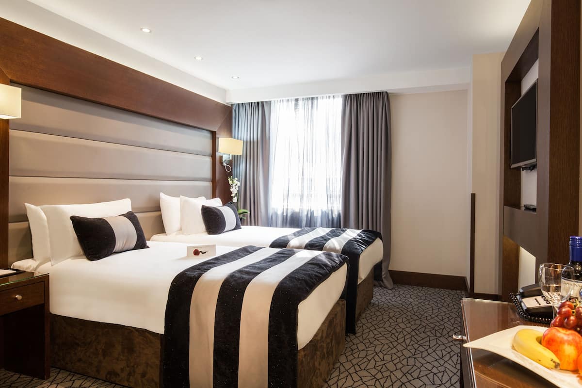 A hotel room with two beds, a TV, a desk with a fruit basket, and a window with curtains.