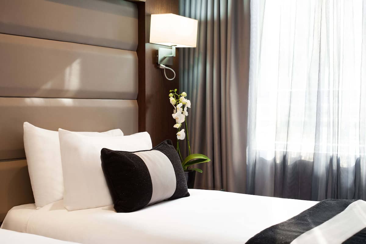 A hotel room with a bed, pillows, a nightstand, a lamp, and a potted plant.