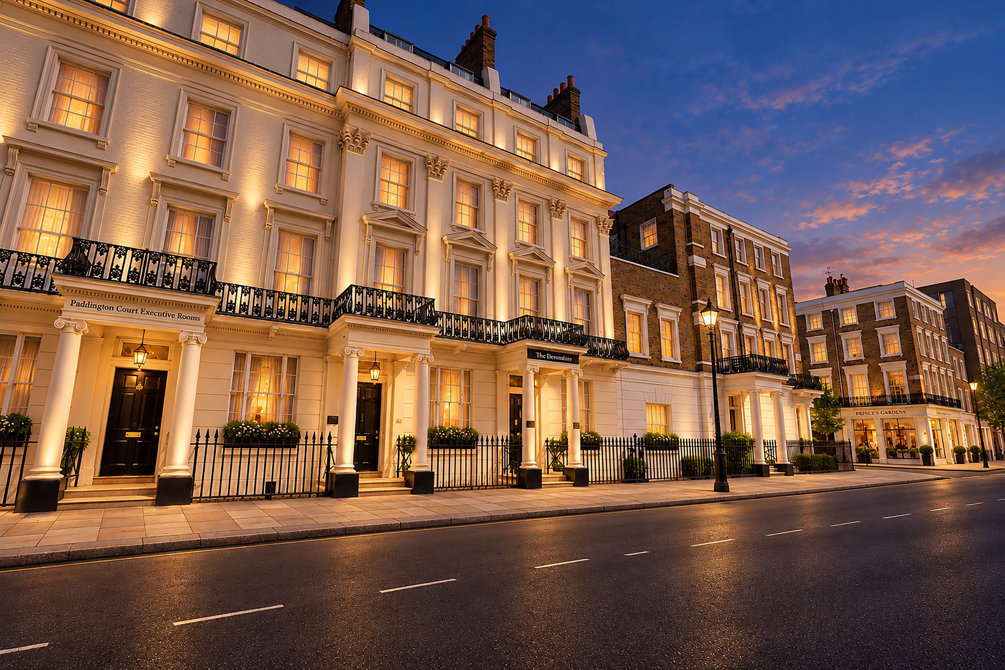 A row of traditional British townhouses with well-lit facades, a clear street, and a calm evening sky.