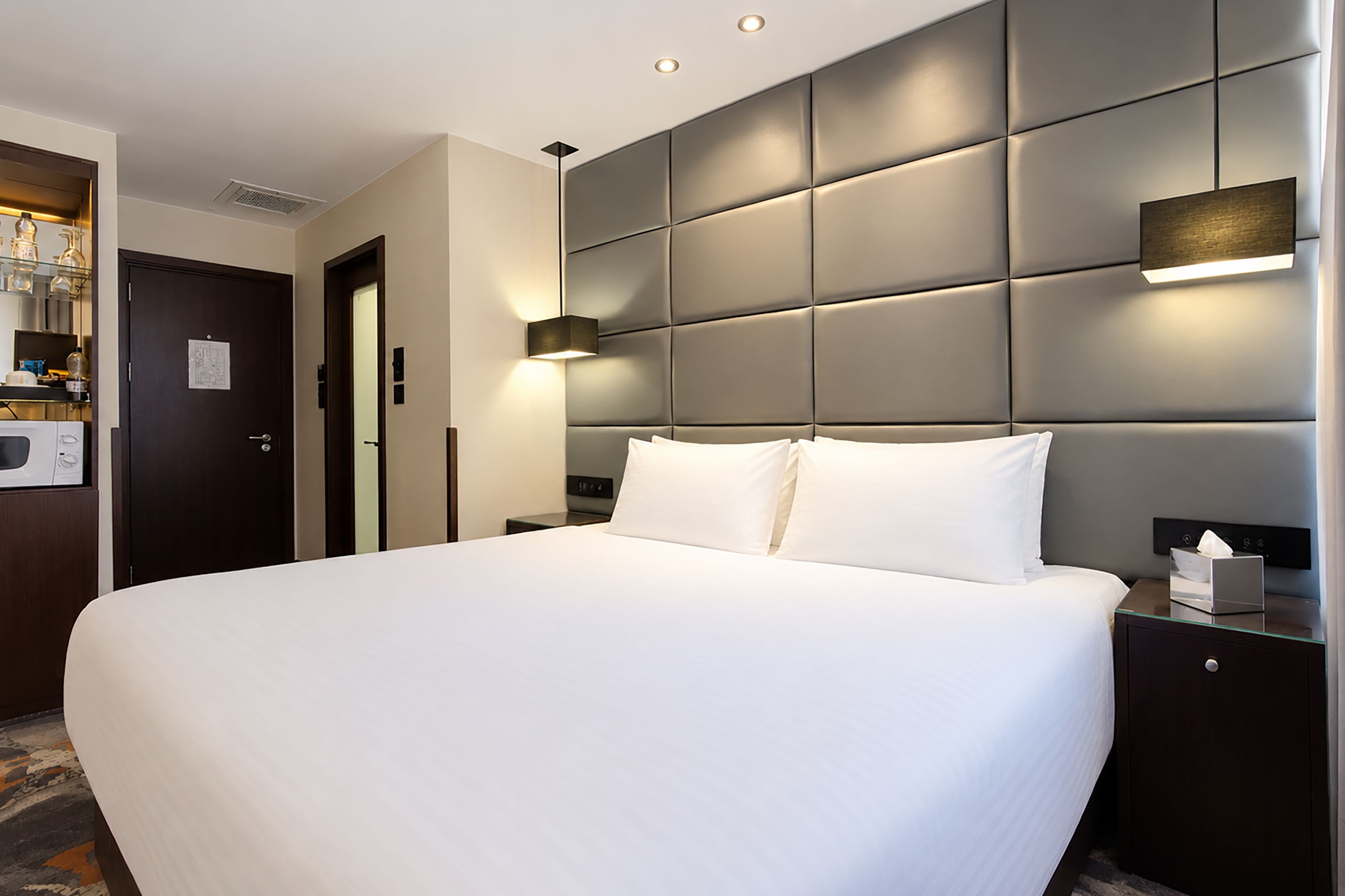 A hotel room with a large bed, a bedside table, a nightstand, a wall-mounted light, and a door.