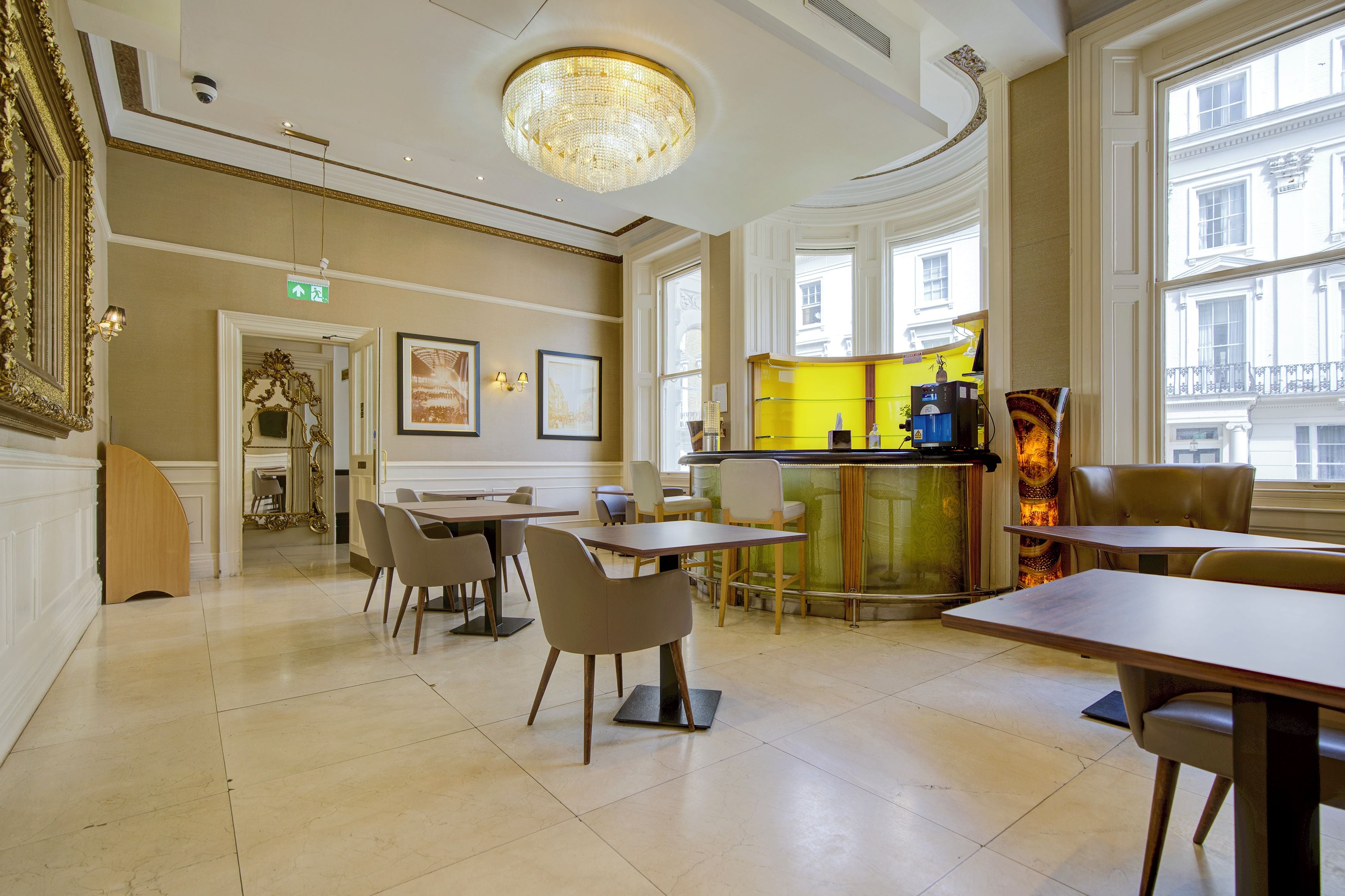 A hotel dining area with tables and chairs, a chandelier, and a view of the street through large windows.
