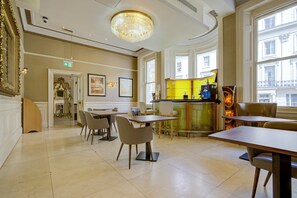 A hotel dining area with tables and chairs, a chandelier, and a view of the street through large windows.
