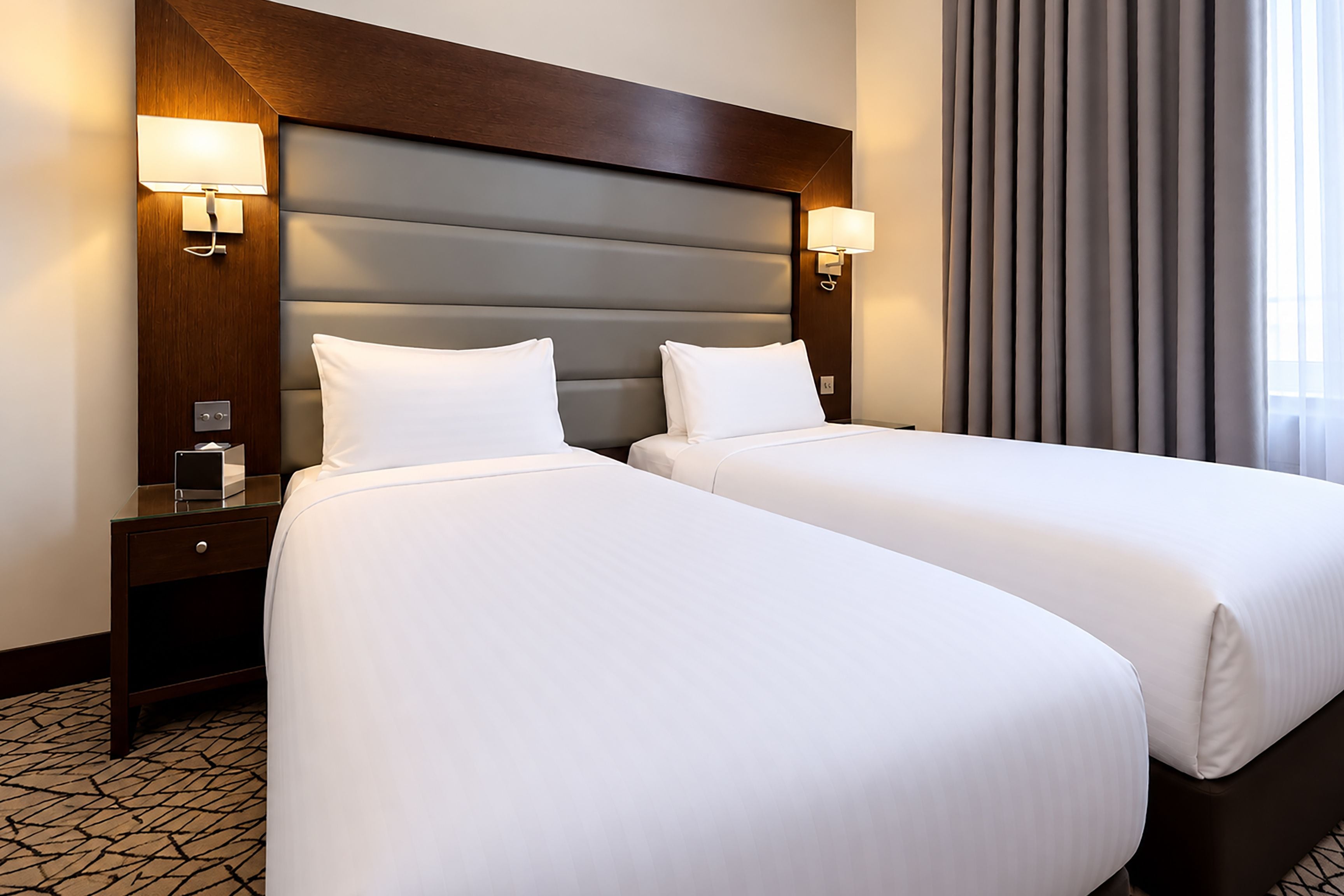 A hotel room with two beds, a wooden headboard, bedside lamps, and a patterned floor.