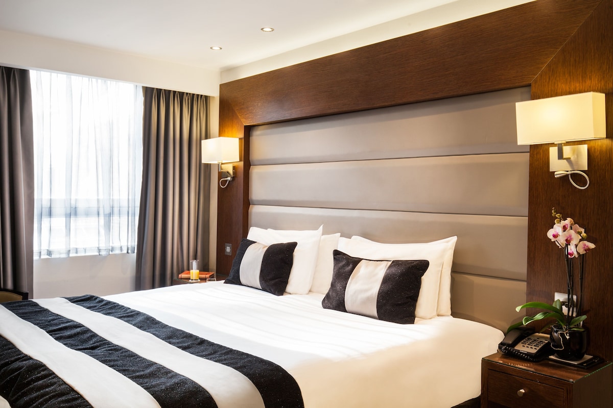 A hotel room with a large bed, two bedside lamps, a nightstand with a phone and a vase of flowers, and a window with curtains.