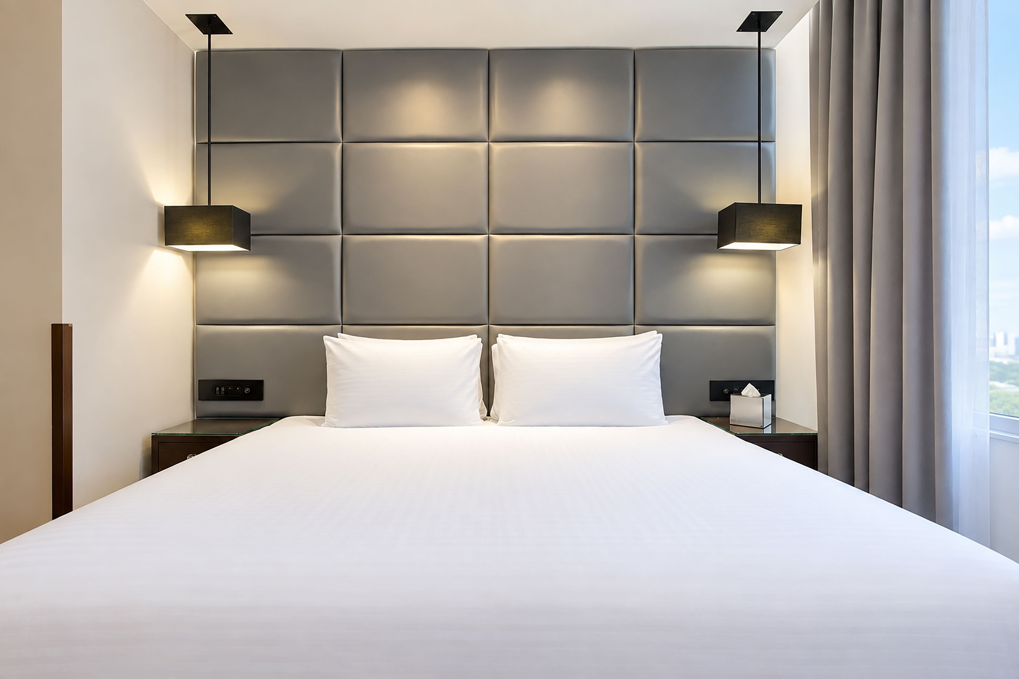 Una habitación de hotel moderna con una cama grande, dos mesitas de noche, un cabecero con iluminación integrada y una ventana con cortinas.