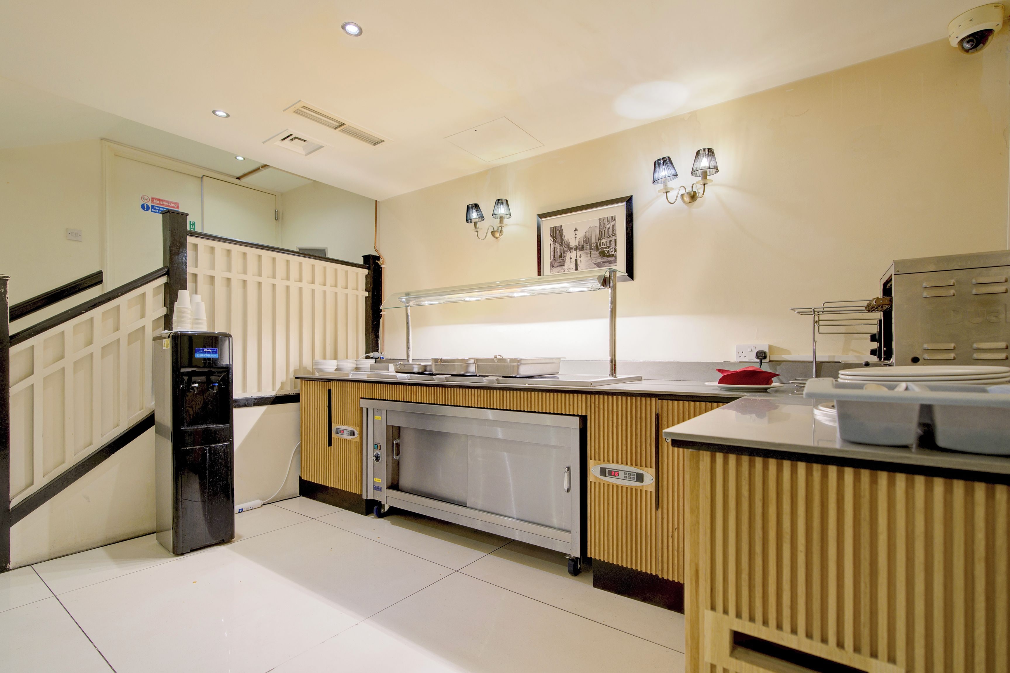 Una cocina moderna con un refrigerador de acero inoxidable, dispensador de agua y microondas. Hay una encimera de madera con un lavamanos y un horno de acero inoxidable. La cocina tiene piso de baldosas y un accesorio de iluminación fijado a la pared.