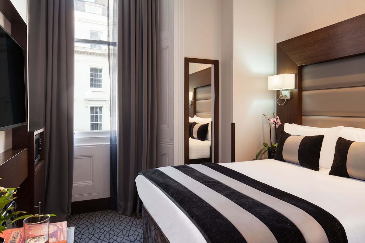 A hotel room with a large bed, a television, a mirror, a bedside lamp, and a view of a building through the window.