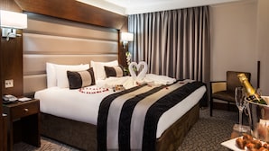 A hotel room with a large bed, a nightstand with a phone, a chair, a table with a bottle of champagne, and a vase with flowers. - Sunday London Paddington Hotel (London)