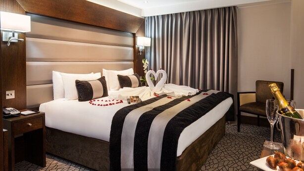 Ein Hotelzimmer mit einem großen Bett, einem Nachttisch mit Telefon, einem Sessel, einem Tisch mit Champagnerflasche und einer Vase mit Blumen.