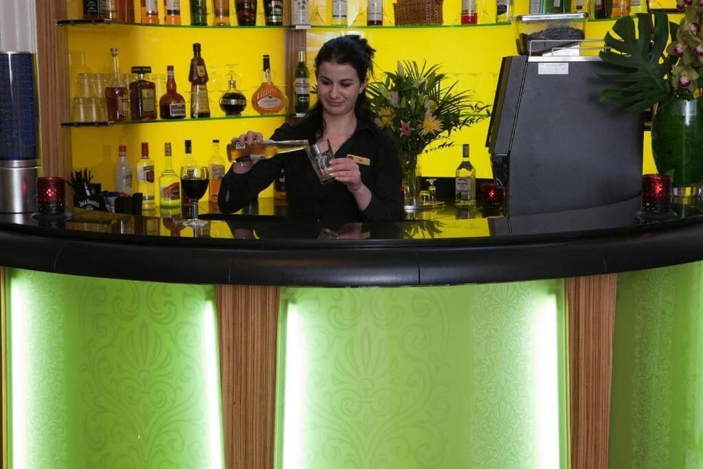 A bar area with a bartender preparing a drink, shelves with various bottles, and a decorative plant.