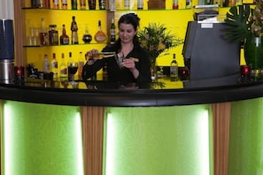 A bar area with a bartender preparing a drink, shelves with various bottles, and a decorative plant. - Sunday London Paddington Hotel (London)