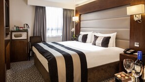 Ein Hotelzimmer mit einem großen Bett, einem Schreibtisch mit Telefon, einer Minibar und einem Fenster mit Vorhängen.