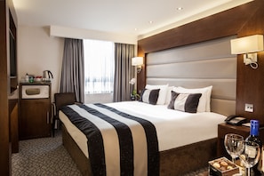 A hotel room with a large bed, a desk with a telephone, a minibar, and a window with curtains.