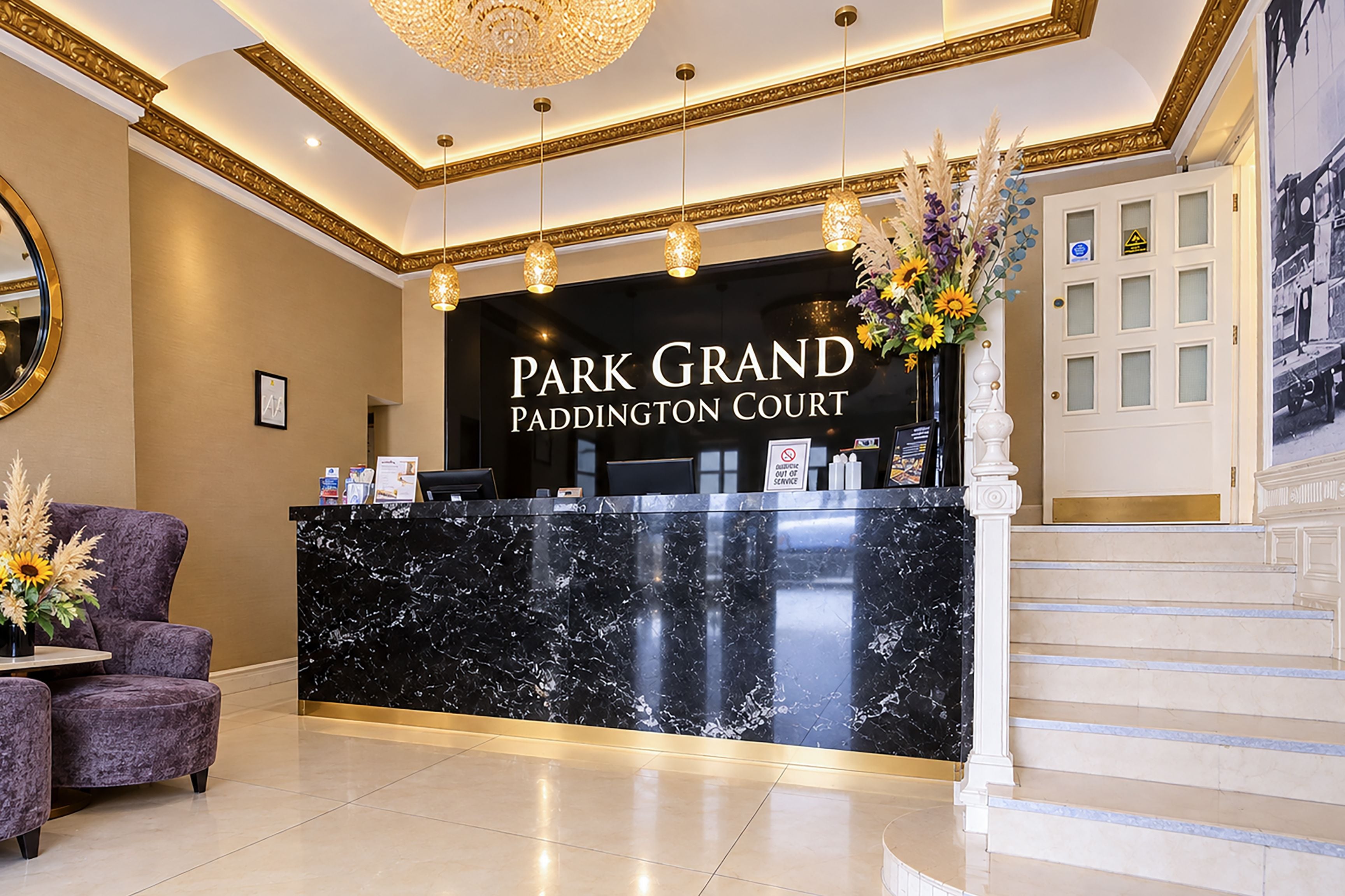A reception desk with a black marble top, a sign reading 'Park Grand Paddington Court,' and a staircase to the right.