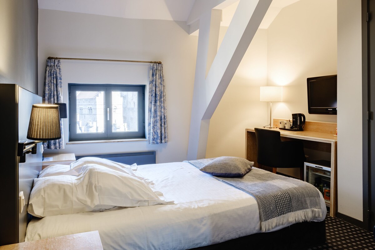Una habitación de hotel con cama, escritorio, televisión y ventana con cortinas.