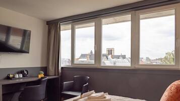 Une chambre d’hôtel équipée d’un lit, d’un bureau, de deux chaises, d’une télévision et d’une grande fenêtre donnant sur la ville.