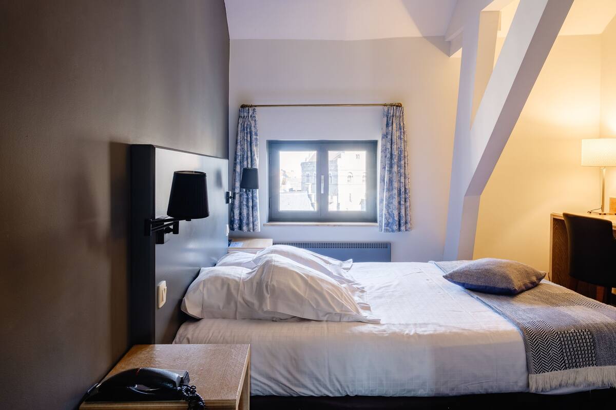 A hotel room with a bed, a bedside table with a lamp, a window with curtains, and a view of the outside.