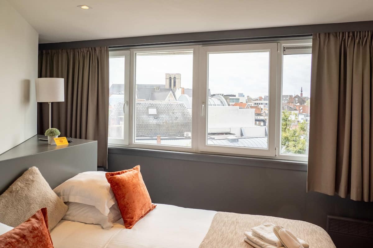 A hotel room with a large window, a bed with pillows, a bedside table with a lamp, and a sign indicating no smoking.