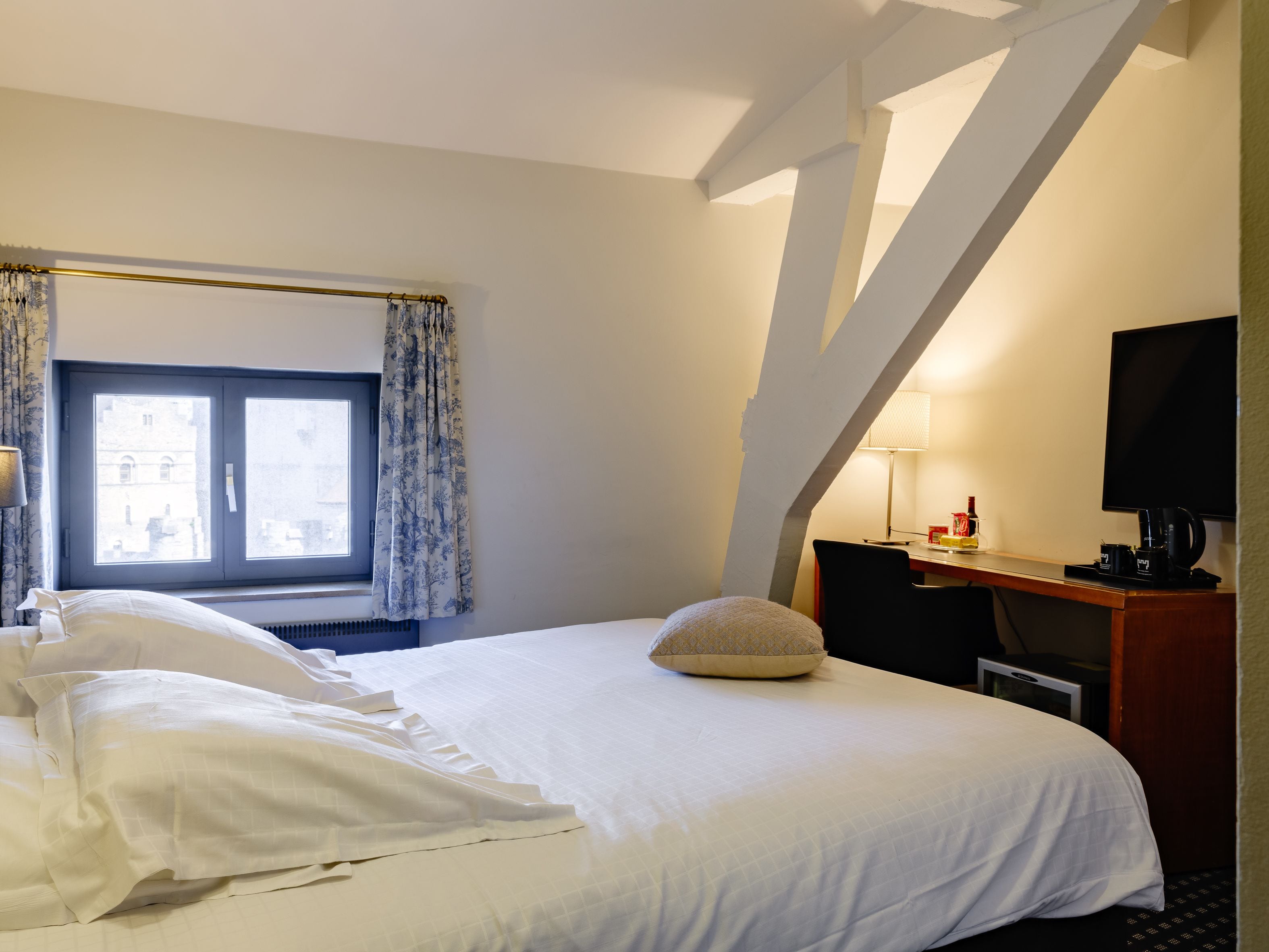 A hotel room with a bed, a desk with a computer, a TV, and a window with curtains.