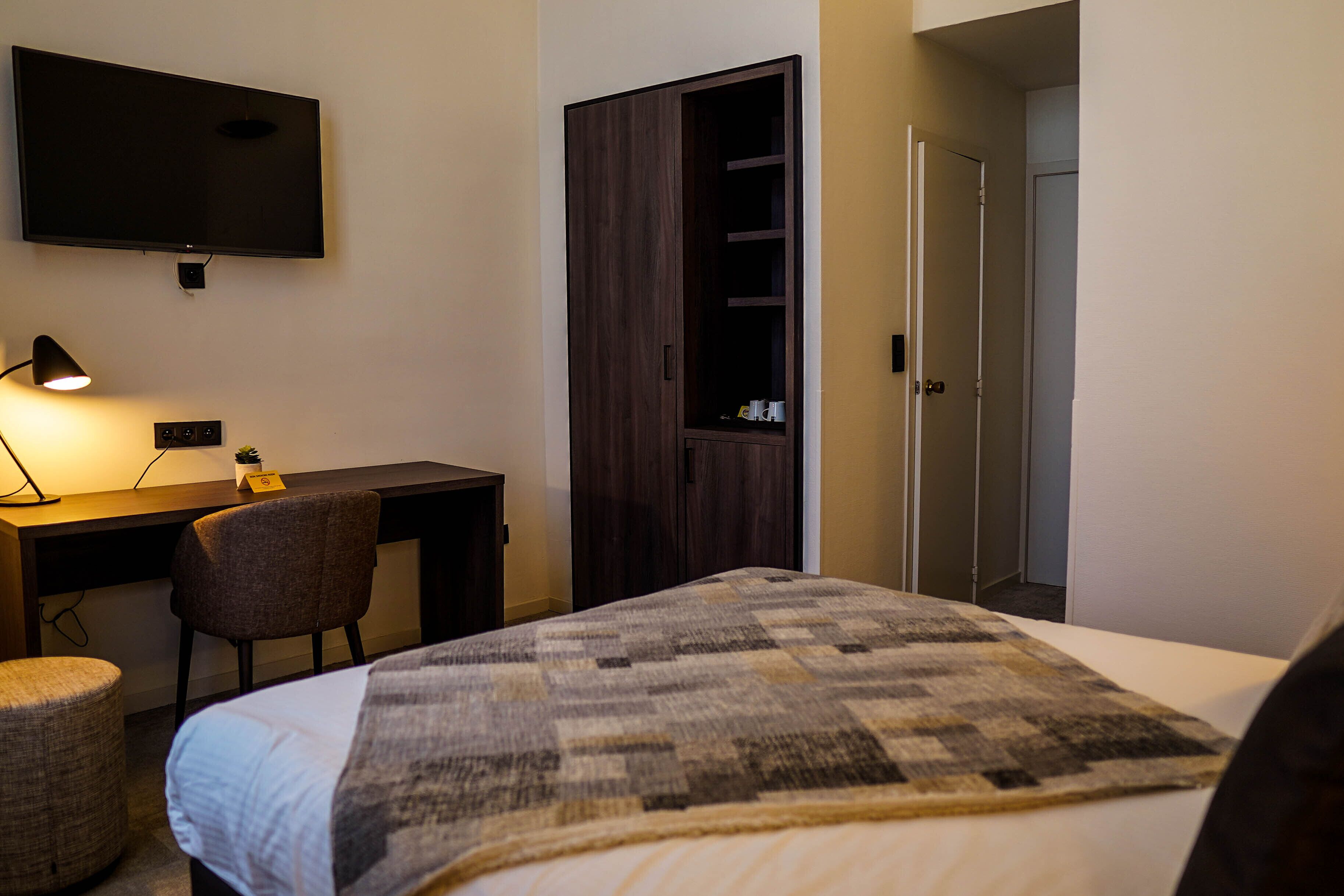 Habitación de hotel con cama, escritorio con silla, televisor montado en la pared y armario.