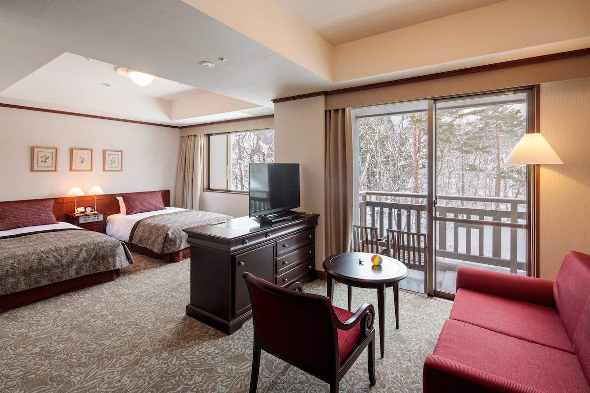 A hotel room with two beds, a TV, a desk, a chair, a red sofa, and large windows with a view of trees.