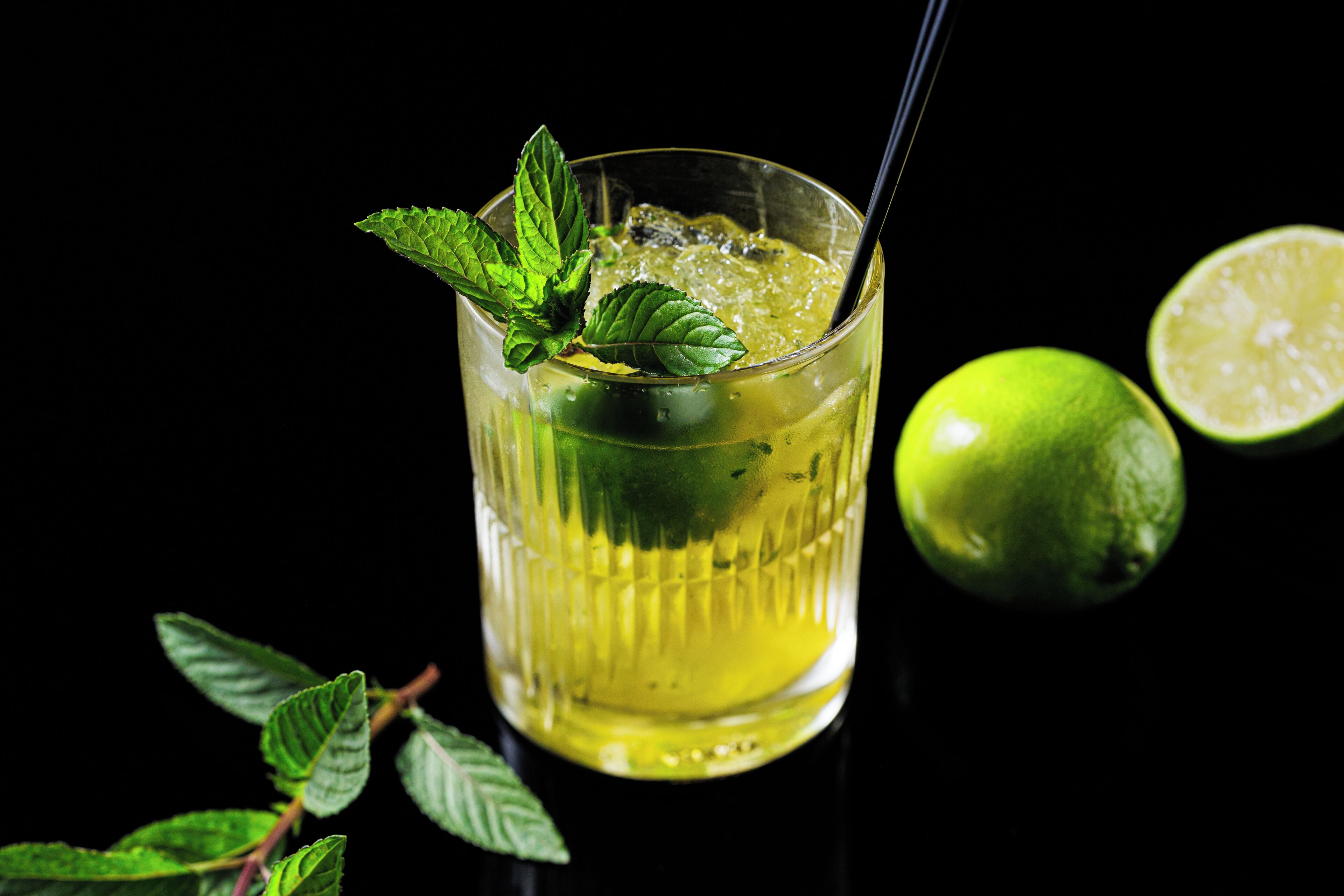 A glass of yellow beverage with mint leaves and a lime half, against a black background.