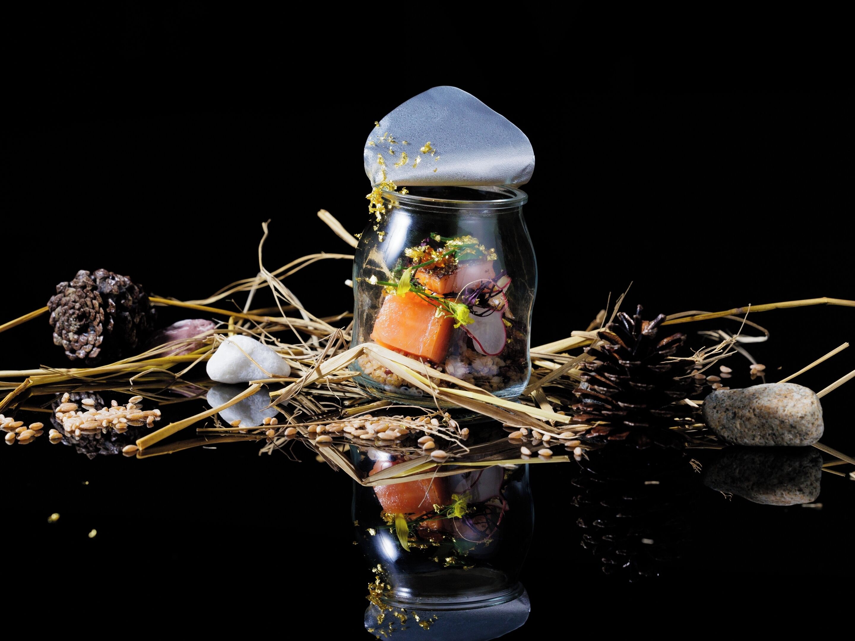 A glass jar with a lid, containing a piece of salmon, radish, and some greenery, surrounded by pine cones, rocks, and straw on a reflective surface.